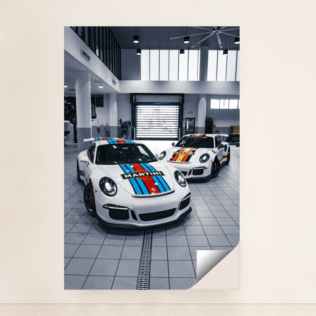 This photo shows two Porsche sports cars with Martini Racing liveries parked in a bright modern garage, unframed.