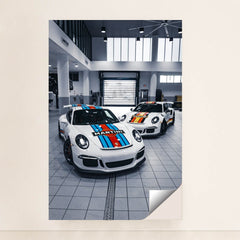 This photo shows two Porsche sports cars with Martini Racing liveries parked in a bright modern garage, unframed.