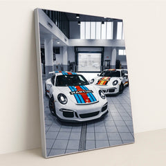 This photo shows two Porsche sports cars with Martini Racing liveries parked in a bright modern garage, in a silver frame.