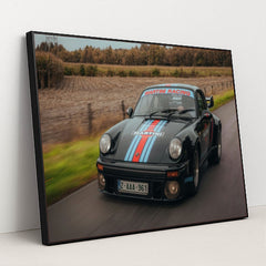 This photo features a vintage black Porsche with Martini Racing stripes driving on a rural road, in a black frame.