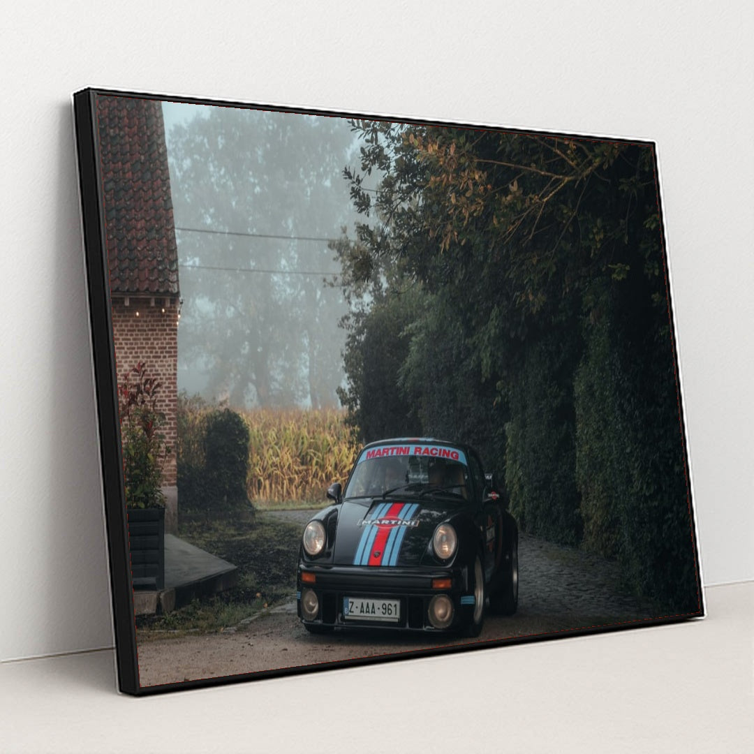 This photo shows a black Porsche with Martini Racing livery parked on a cobblestone driveway next to a brick house on a foggy morning, in a black frame.