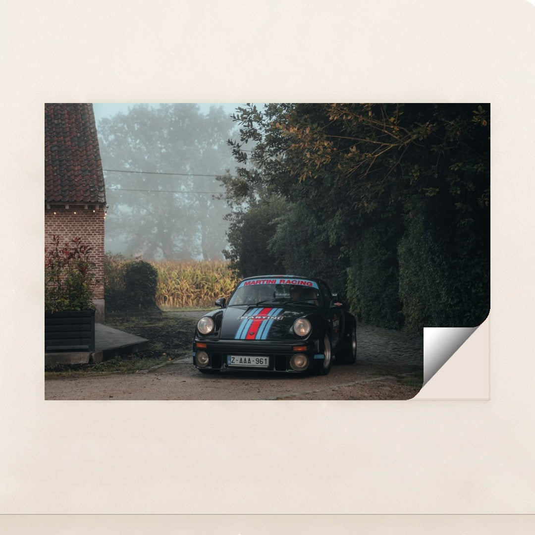 This photo shows a black Porsche with Martini Racing livery parked on a cobblestone driveway next to a brick house on a foggy morning, unframed.