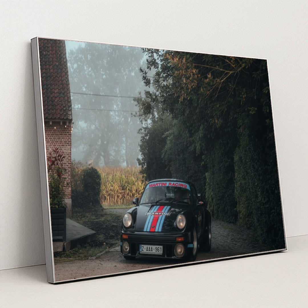 This photo shows a black Porsche with Martini Racing livery parked on a cobblestone driveway next to a brick house on a foggy morning, in a silver frame.