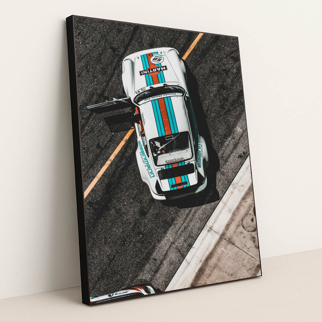 This photo shows an overhead view of a white Porsche race car with Martini stripes and an open door on a racetrack, in a black frame.