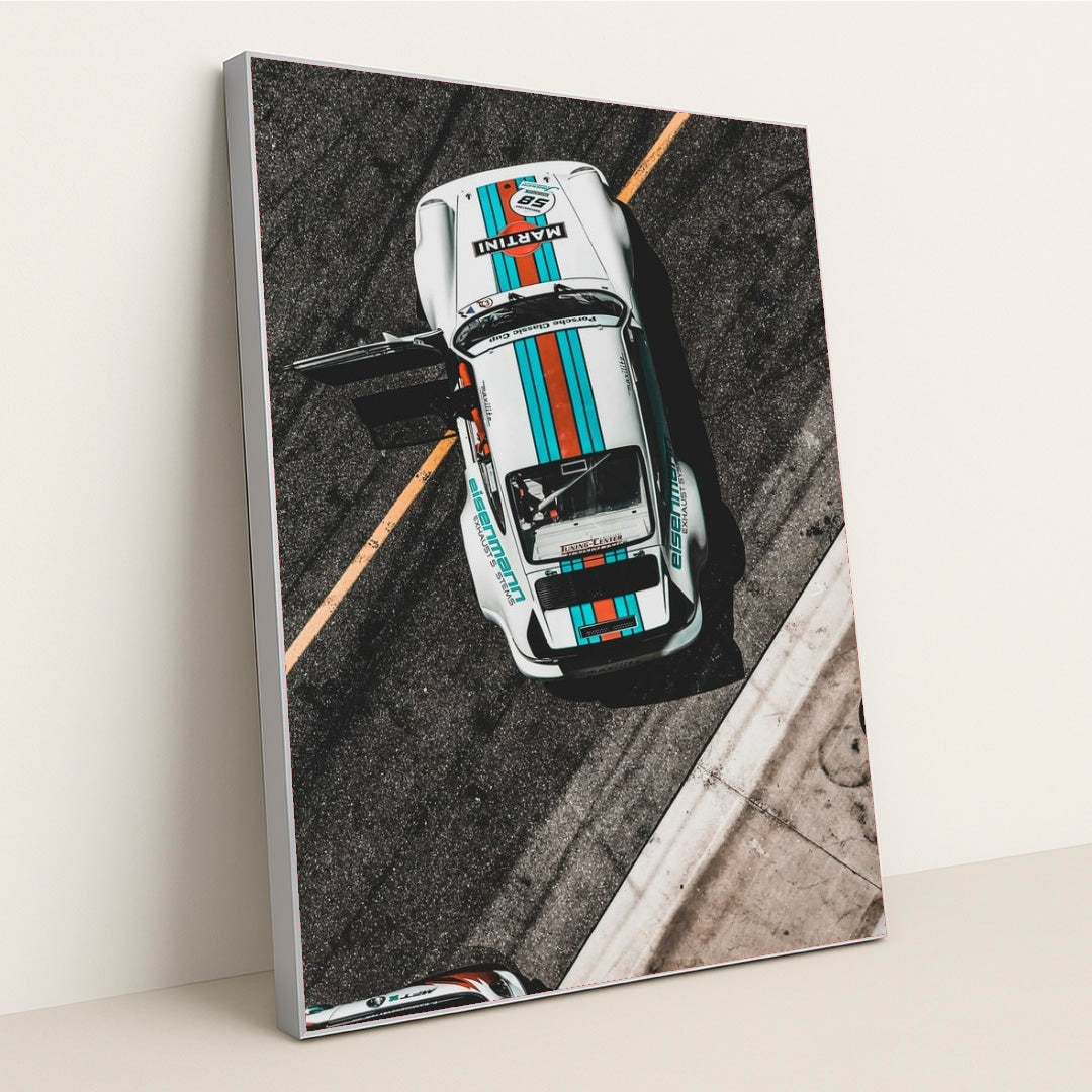 This photo shows an overhead view of a white Porsche race car with Martini stripes and an open door on a racetrack, in a silver frame.