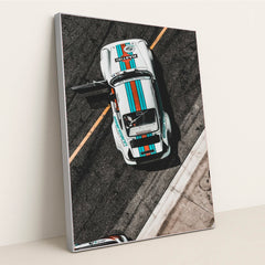 This photo shows an overhead view of a white Porsche race car with Martini stripes and an open door on a racetrack, in a silver frame.