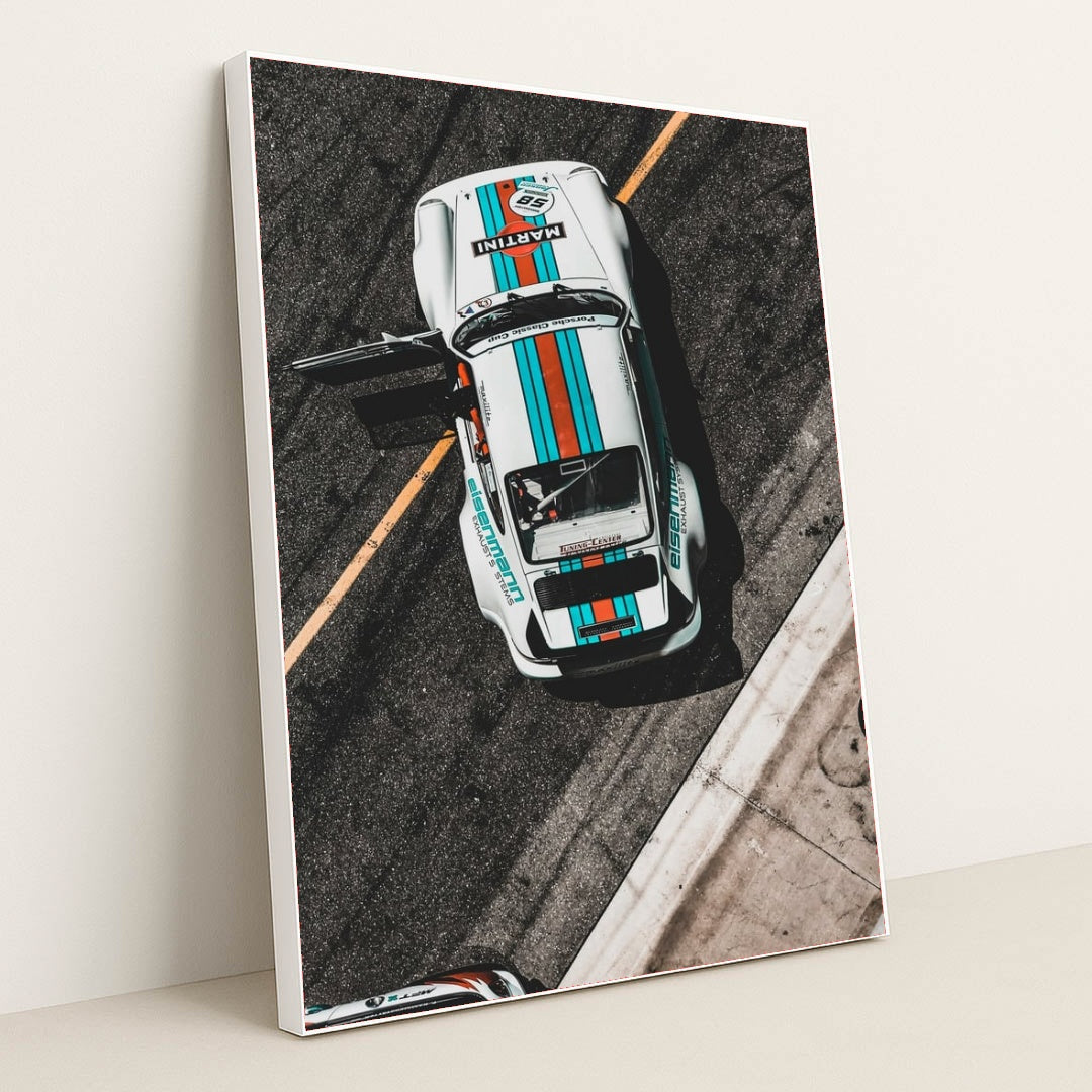 This photo shows an overhead view of a white Porsche race car with Martini stripes and an open door on a racetrack, in a white frame.