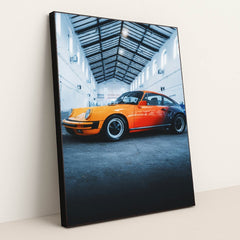 This photo features an orange and red classic Porsche sports car parked inside a tall industrial warehouse, in a black frame.
