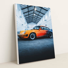 This photo features an orange and red classic Porsche sports car parked inside a tall industrial warehouse, in a white frame.