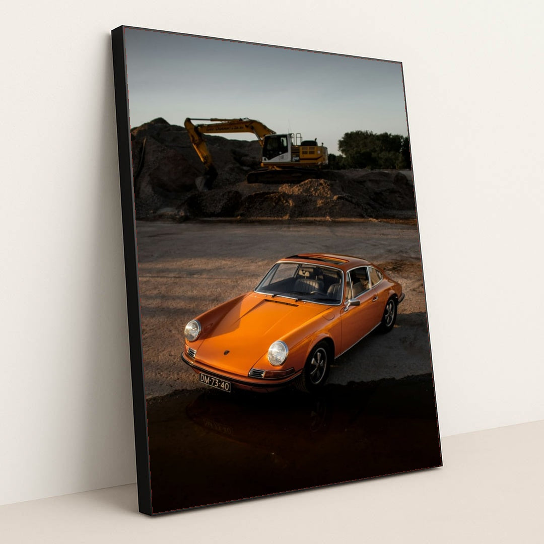 This photo shows an orange classic Porsche 911 parked on gravel at sunset with a yellow excavator in the background, in a black frame.