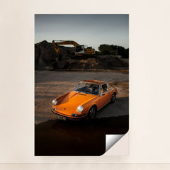 This photo shows an orange classic Porsche 911 parked on gravel at sunset with a yellow excavator in the background, minimalist unframed style.