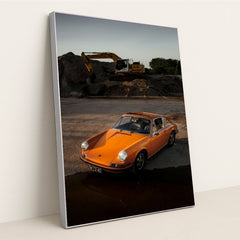This photo shows an orange classic Porsche 911 parked on gravel at sunset with a yellow excavator in the background, in a silver frame.