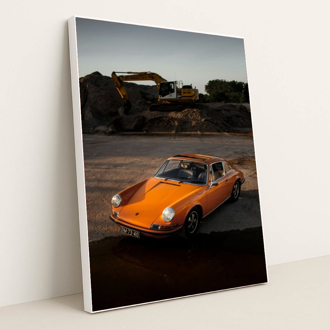 This photo shows an orange classic Porsche 911 parked on gravel at sunset with a yellow excavator in the background, in a white frame.