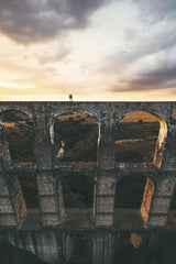 Mikhail Nilov - A lone figure stands atop a historic aqueduct at sunset, surrounded by dramatic clouds.