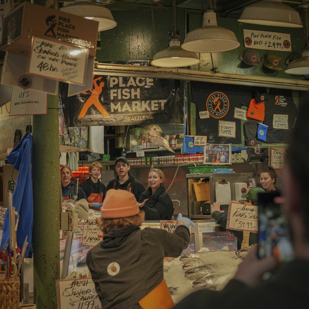 Eddie Ortiz - Lively atmosphere of Pike Place Fish Market with people engaged in fish tossing and sales activities.