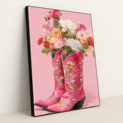 This artwork shows two pink floral cowboy boots filled with colorful roses and flowers against a solid pink background, in a black frame.