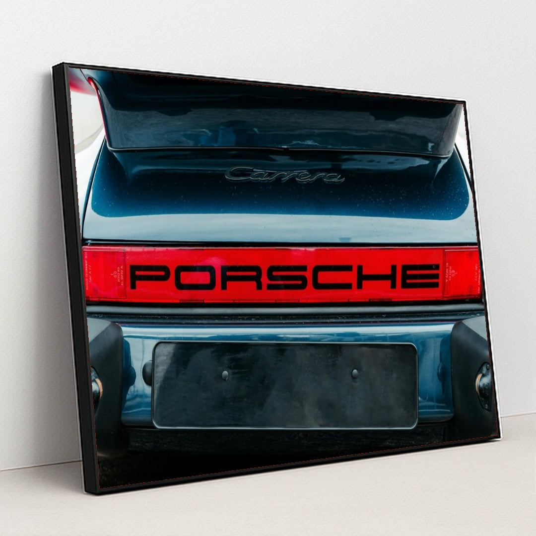 This photo shows the rear of a classic Porsche Carrera with a bold red Porsche logo and open trunk, in a black frame.