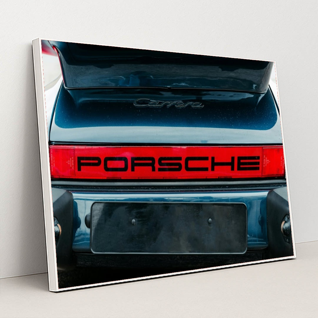 This photo shows the rear of a classic Porsche Carrera with a bold red Porsche logo and open trunk, in a white frame.