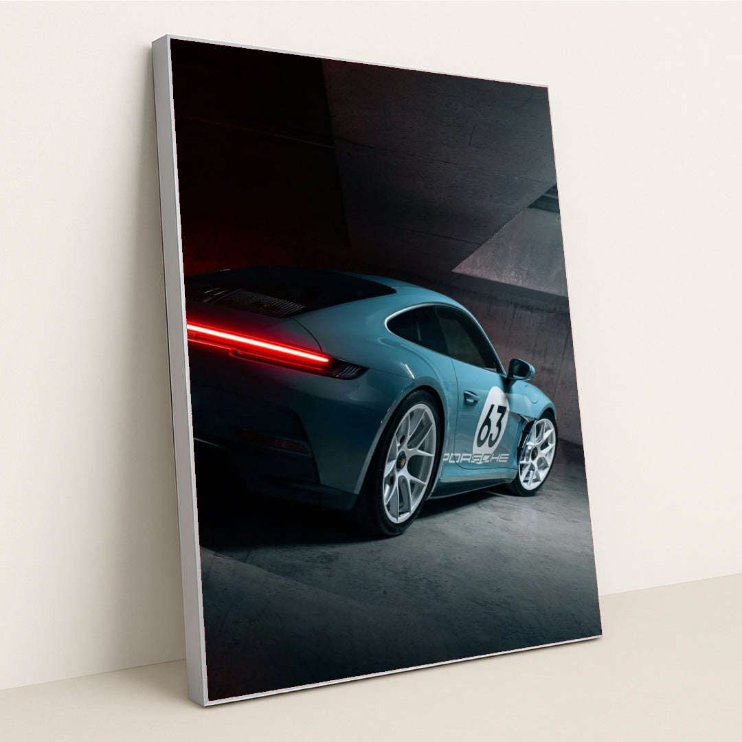 This photo shows a silver Porsche sports car with number 63 and glowing taillights in a modern concrete garage, in a silver frame.