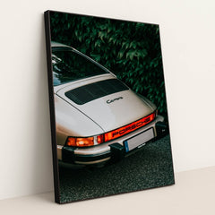 This photo shows the rear end of a classic silver Porsche Carrera parked in front of green leafy foliage, in a black frame.