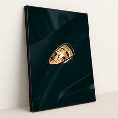 This photo shows a close-up of a classic gold and black Porsche crest badge on a dark car hood, in a black frame.