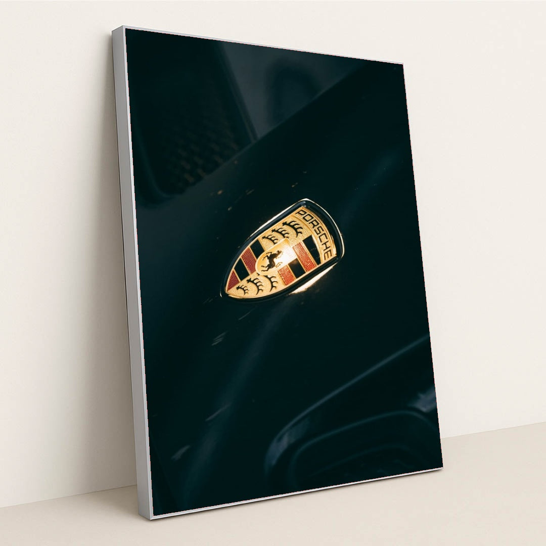 This photo shows a close-up of a classic gold and black Porsche crest badge on a dark car hood, in a silver frame.
