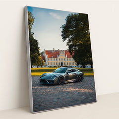 This photo shows a silver Porsche sports car parked on a cobblestone driveway in front of a large historic mansion, in a silver frame.