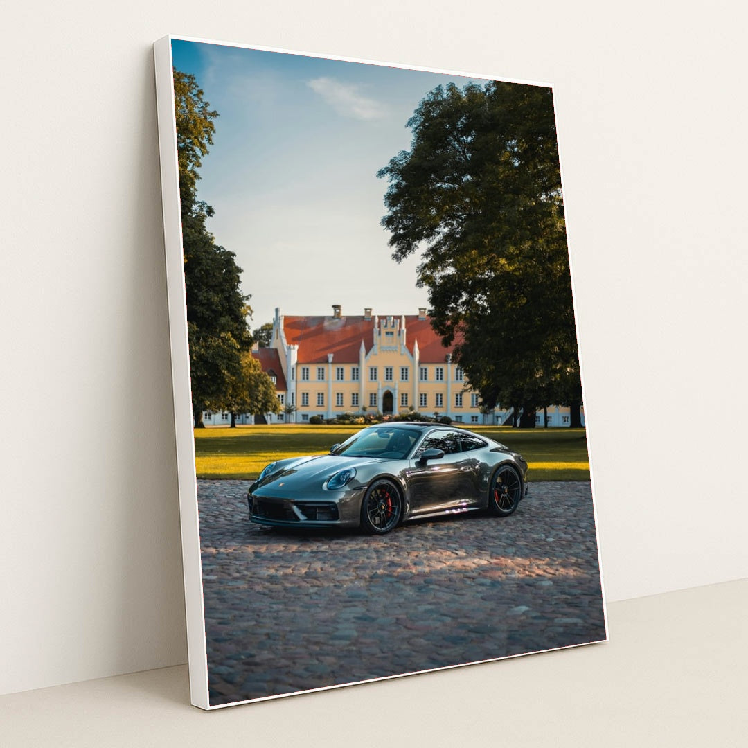 This photo shows a silver Porsche sports car parked on a cobblestone driveway in front of a large historic mansion, in a white frame.