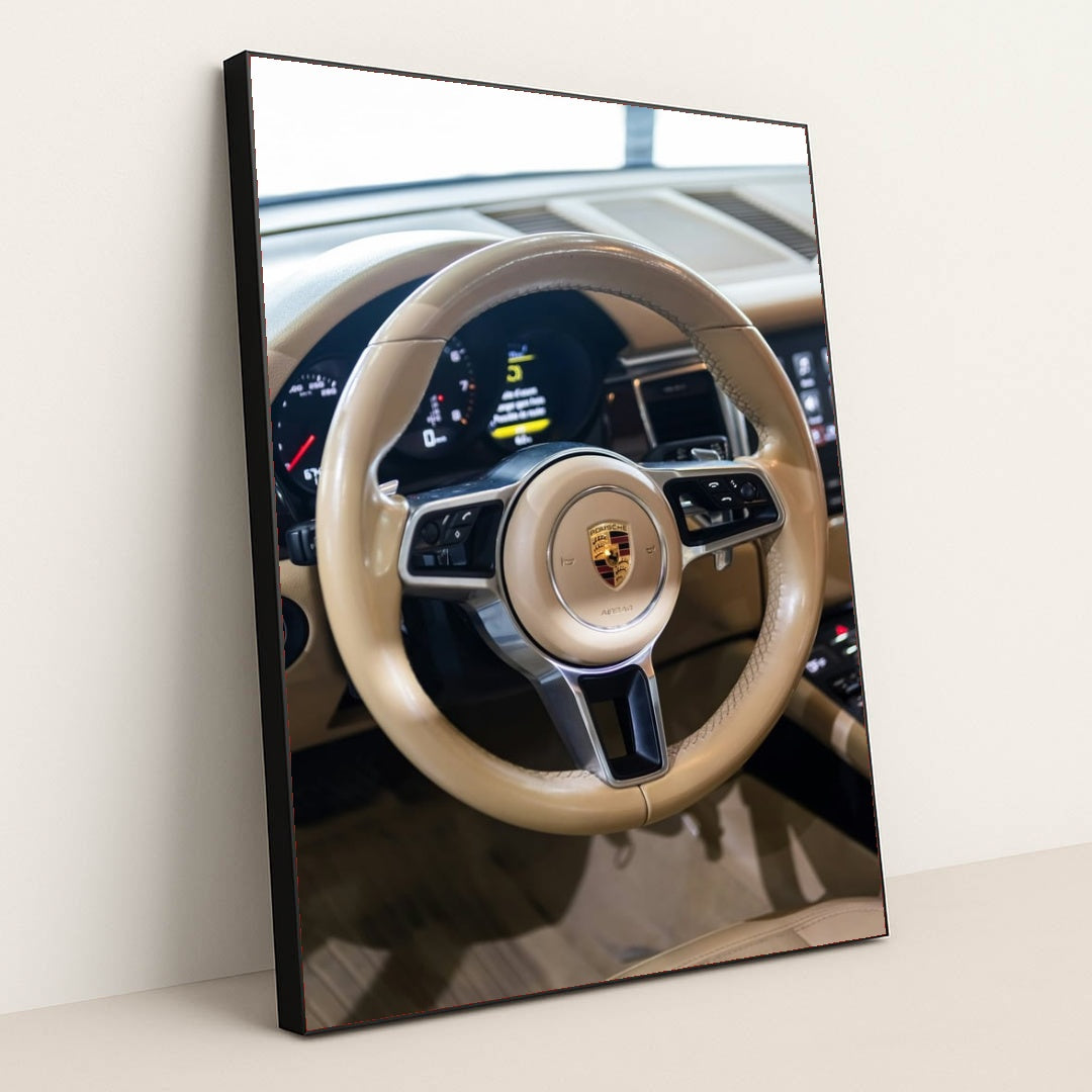 This photo shows the tan leather steering wheel and dashboard of a Porsche with the crest emblem in the center, in a black frame.