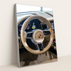 This photo shows the tan leather steering wheel and dashboard of a Porsche with the crest emblem in the center, in a silver frame.