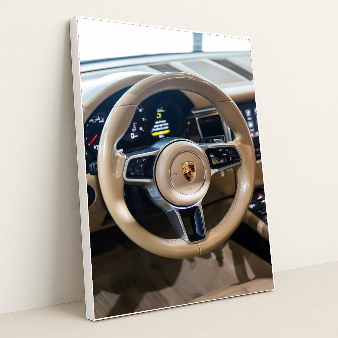 This photo shows the tan leather steering wheel and dashboard of a Porsche with the crest emblem in the center, in a white frame.