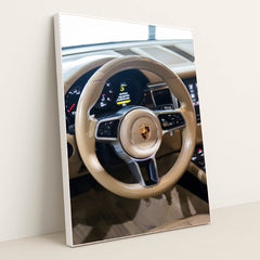This photo shows the tan leather steering wheel and dashboard of a Porsche with the crest emblem in the center, in a white frame.