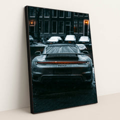 This photo shows the rear view of a silver Porsche sports car covered in raindrops on a city street at night, in a black frame.