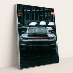 This photo shows the rear view of a silver Porsche sports car covered in raindrops on a city street at night, in a silver frame.