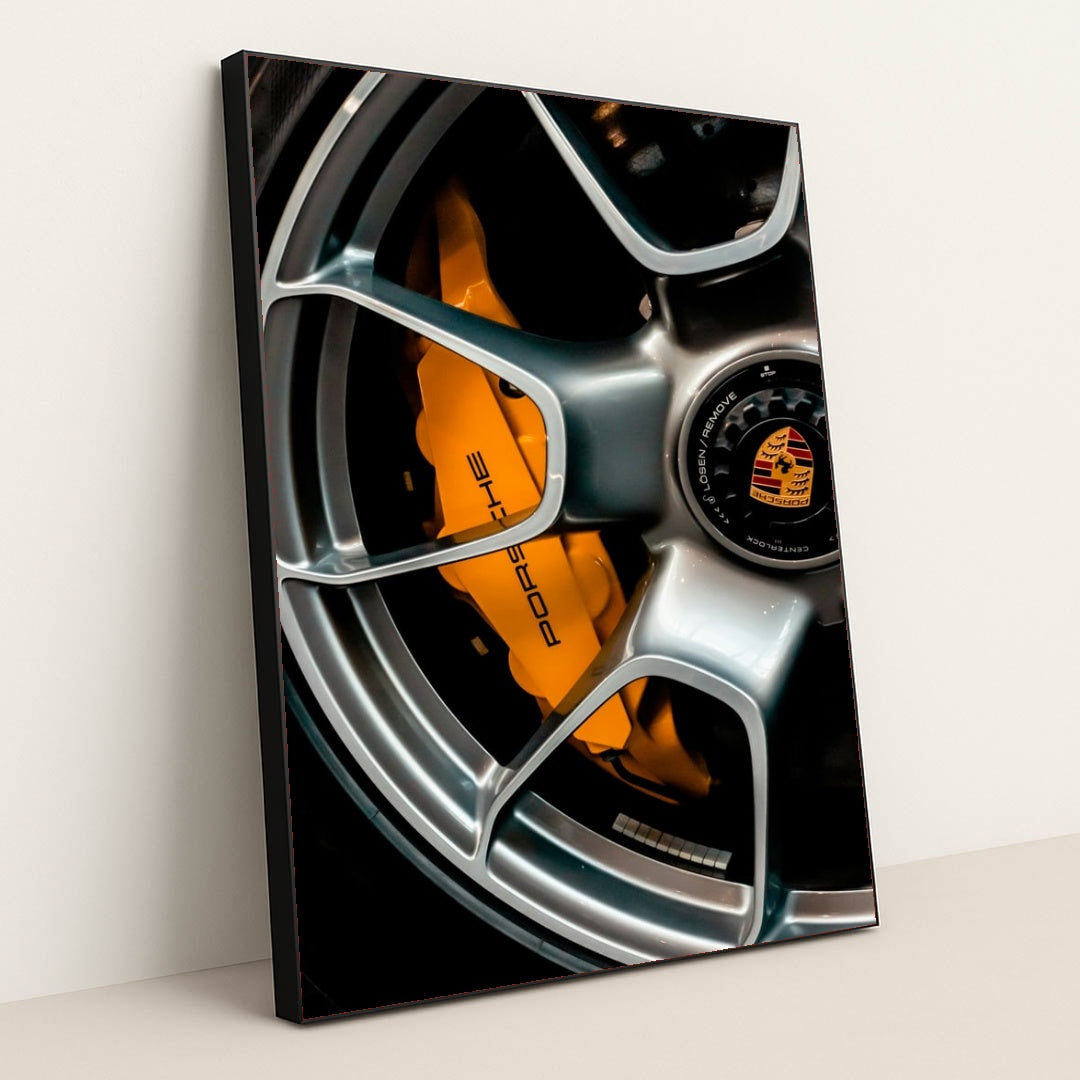 This photo shows a close-up of a silver Porsche wheel with an orange brake caliper and crest logo, in a black frame.