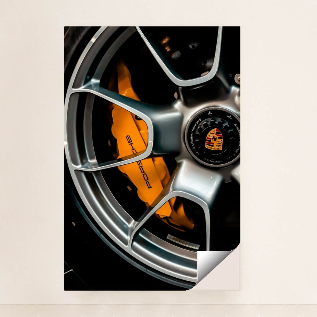 This photo shows a close-up of a silver Porsche wheel with an orange brake caliper and crest logo, unframed.