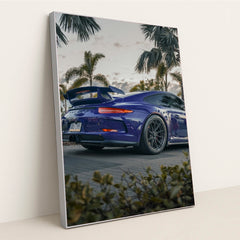 This photo shows a blue Porsche sports car with a large rear wing parked among palm trees at dusk, in a silver frame.