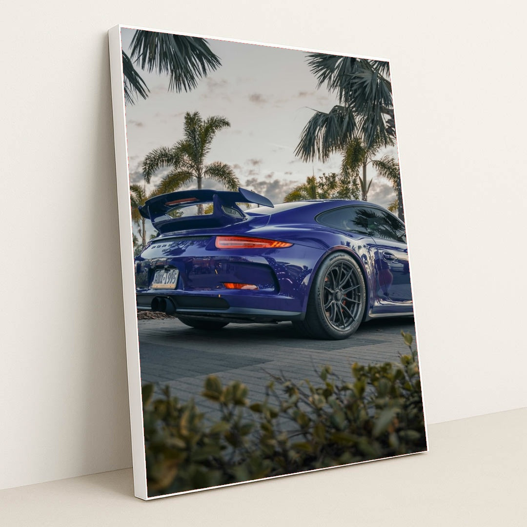 This photo shows a blue Porsche sports car with a large rear wing parked among palm trees at dusk, in a white frame.