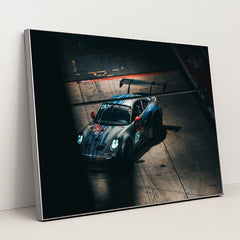 This photo shows a silver Porsche race car with blue stripes and a large rear wing in a dark garage, in a silver frame.