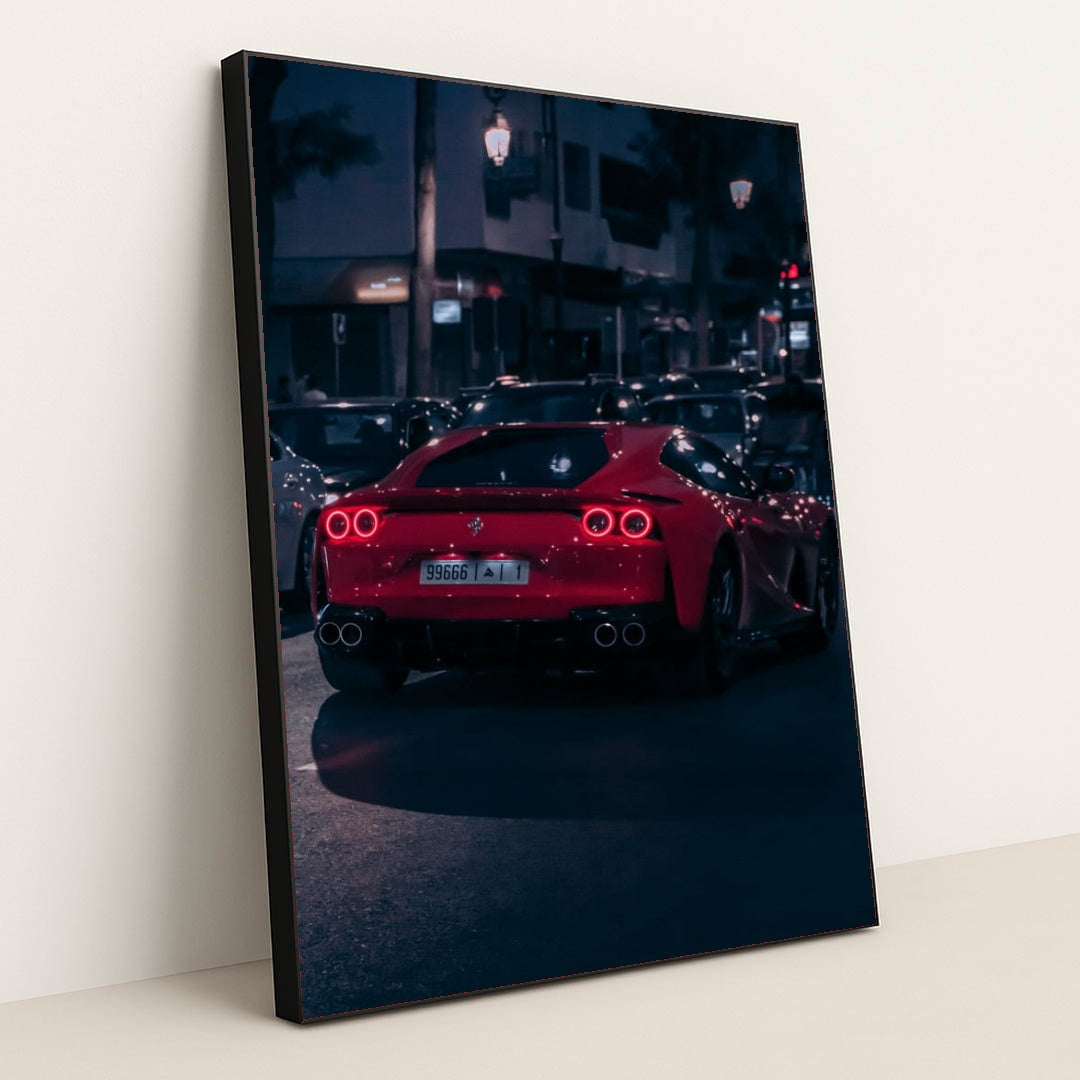 This photo shows a red Ferrari sports car driving at night through city traffic under streetlights, in a black frame.