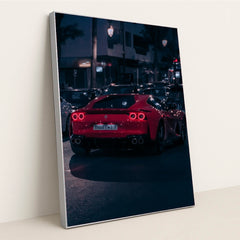 This photo shows a red Ferrari sports car driving at night through city traffic under streetlights, in a silver frame.