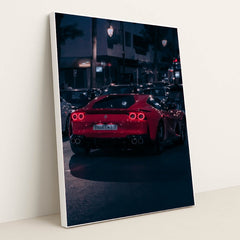This photo shows a red Ferrari sports car driving at night through city traffic under streetlights, in a white frame.