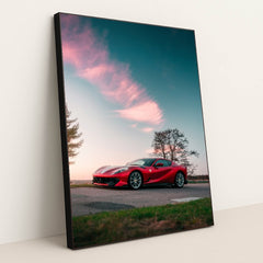 This photo shows a red Ferrari sports car parked on a rural road under a pink-clouded sunset sky, in a black frame.