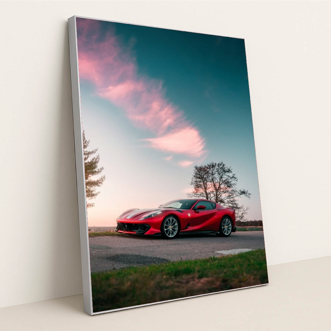 This photo shows a red Ferrari sports car parked on a rural road under a pink-clouded sunset sky, in a silver frame.