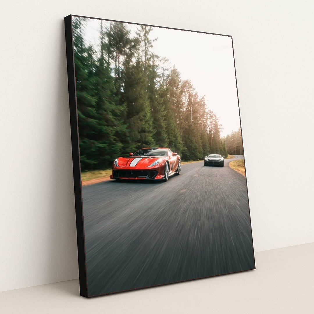 This photo shows a red Ferrari sports car with white stripes leading another car on a winding forest road, in a black frame.