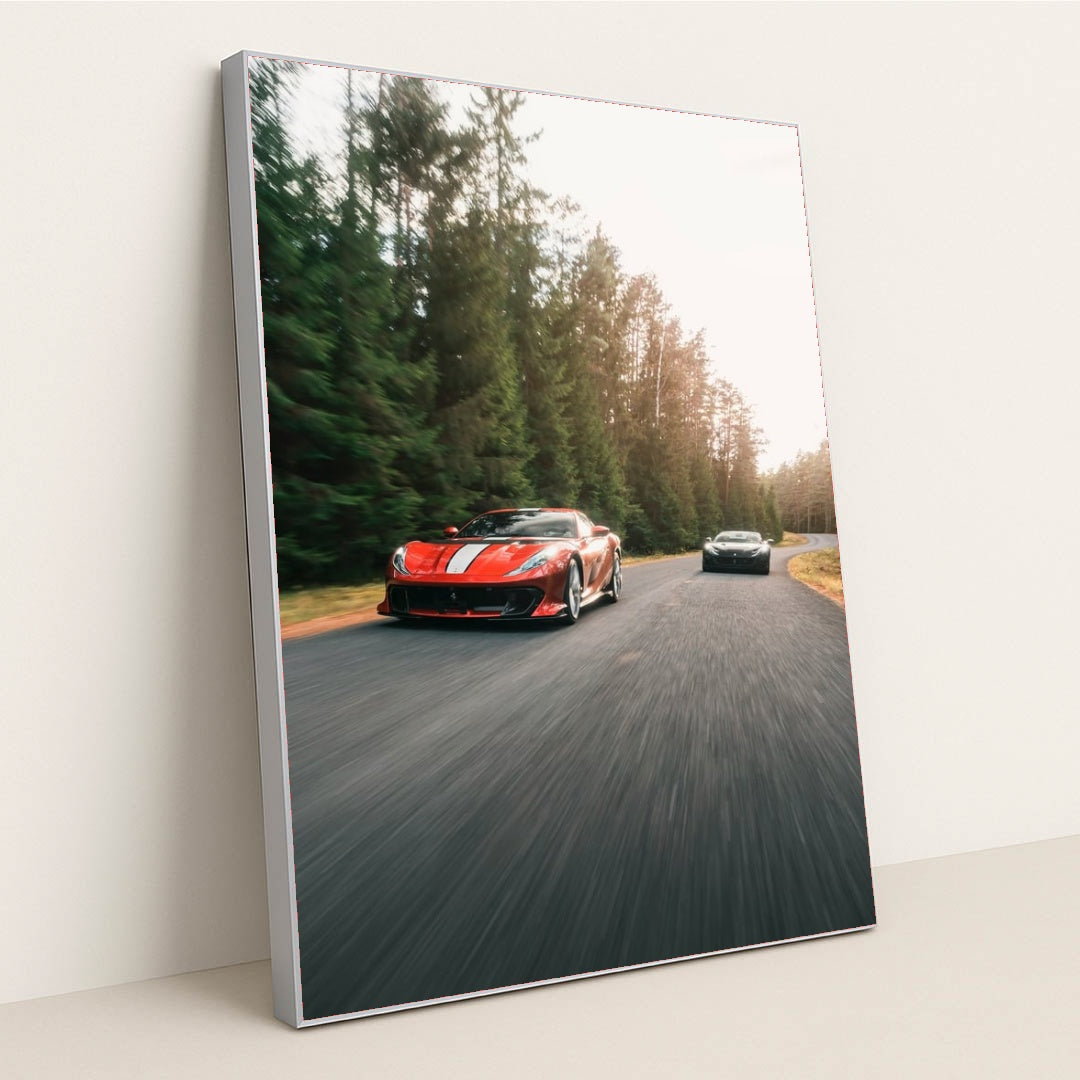 This photo shows a red Ferrari sports car with white stripes leading another car on a winding forest road, in a silver frame.