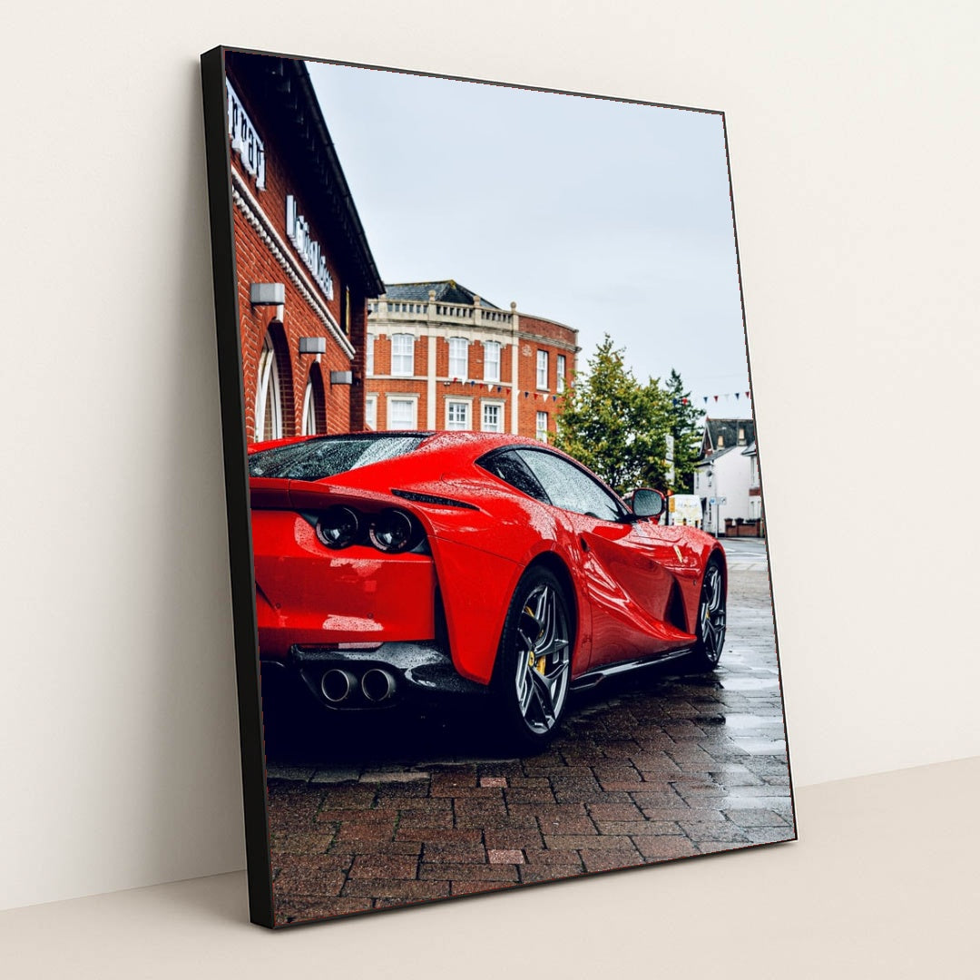 This photo shows the rear three-quarter view of a red Ferrari 812 Superfast parked on a wet city street, in a black frame.