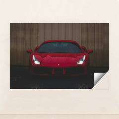 This photo shows the front view of a red Ferrari sports car with headlights on, parked on wet pavement near a metal wall, minimalist unframed style.