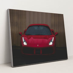 This photo shows the front view of a red Ferrari sports car with headlights on, parked on wet pavement near a metal wall, in a silver frame.