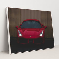 This photo shows the front view of a red Ferrari sports car with headlights on, parked on wet pavement near a metal wall, in a white frame.